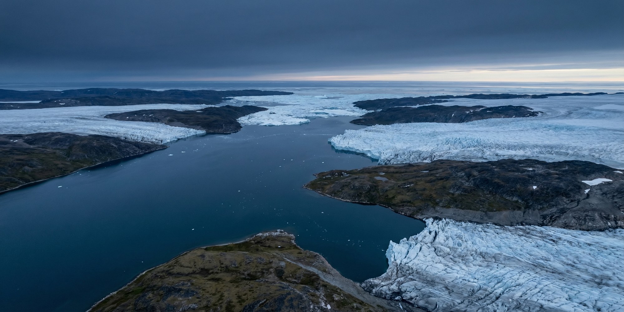 Aerial view of Greenland's Arctic coastline at twilight — dark fjord waters, blue-white glacial ice formations, and remote tundra under a pale sky