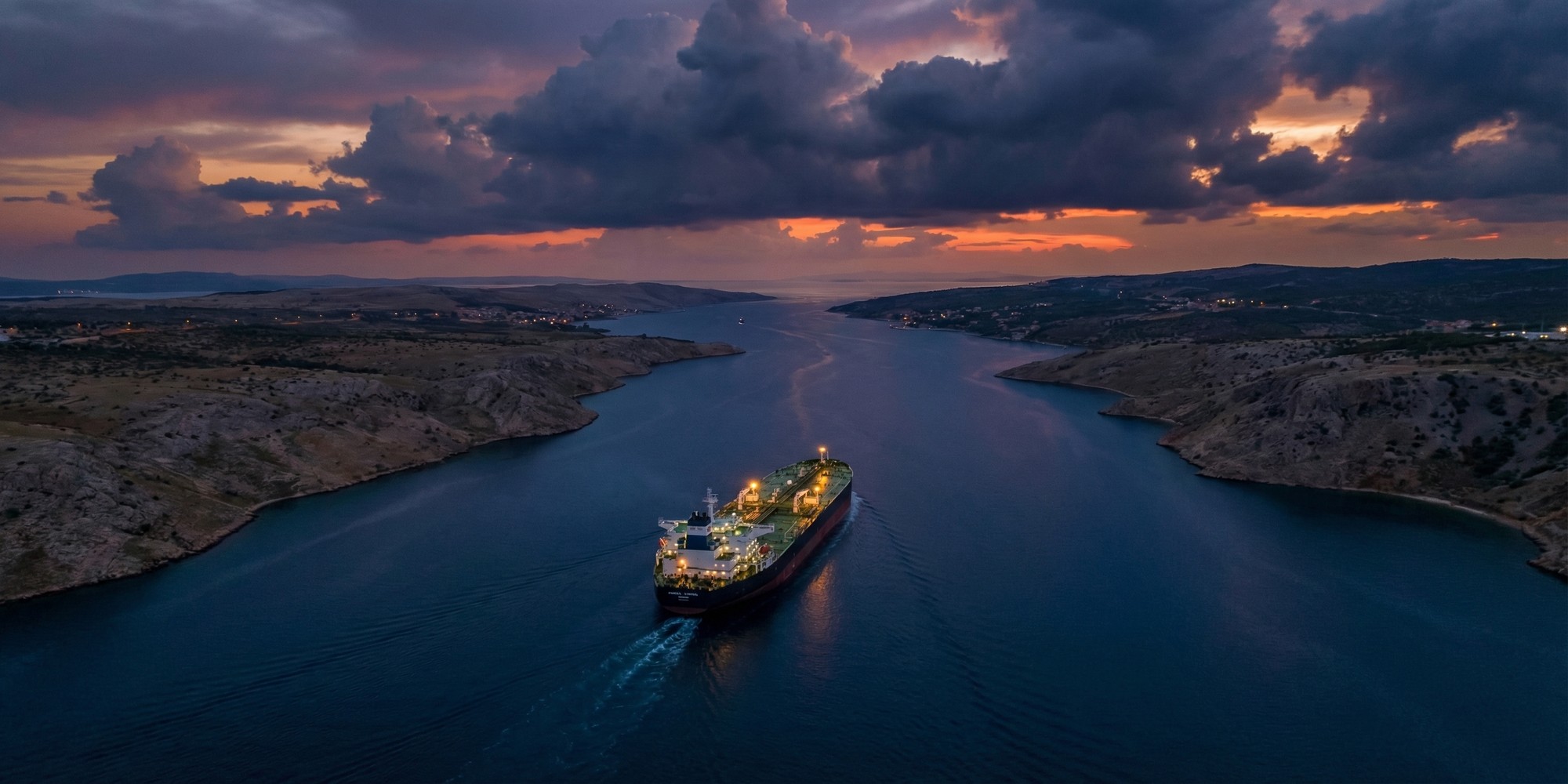 Aerial view of a large oil tanker navigating a strategic maritime strait at dusk, with dramatic cloud formations and distant coastlines on both sides