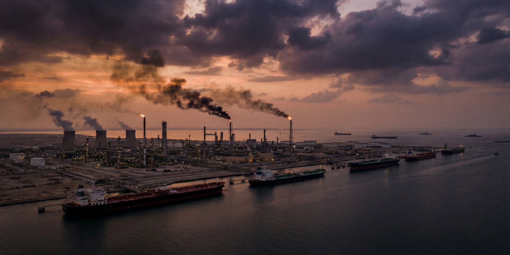 Persian Gulf oil refinery infrastructure with supertankers in the waterway below a storm-laden sky, symbolizing the disruption to Gulf energy flows